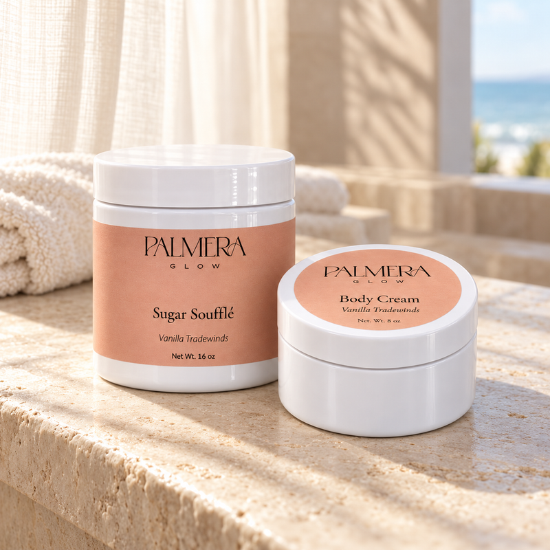 Vanilla Tradewinds Glow Duo: 16 oz Sugar Soufflé and 8 oz Body Cream on sunlit stone beside a towel and an ocean view.